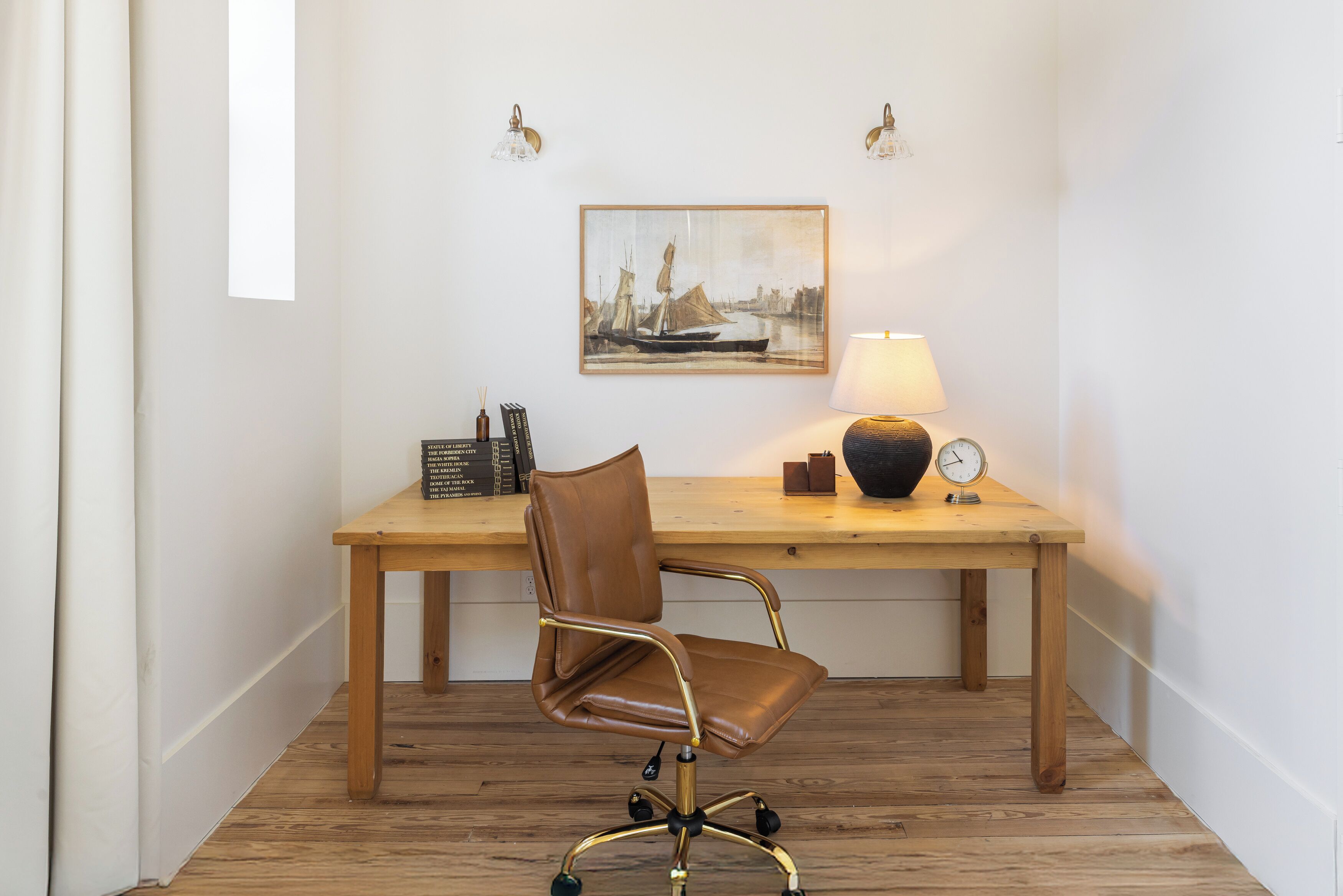 A wooden desk with a brown leather chair, a framed painting, a lamp, and a clock.