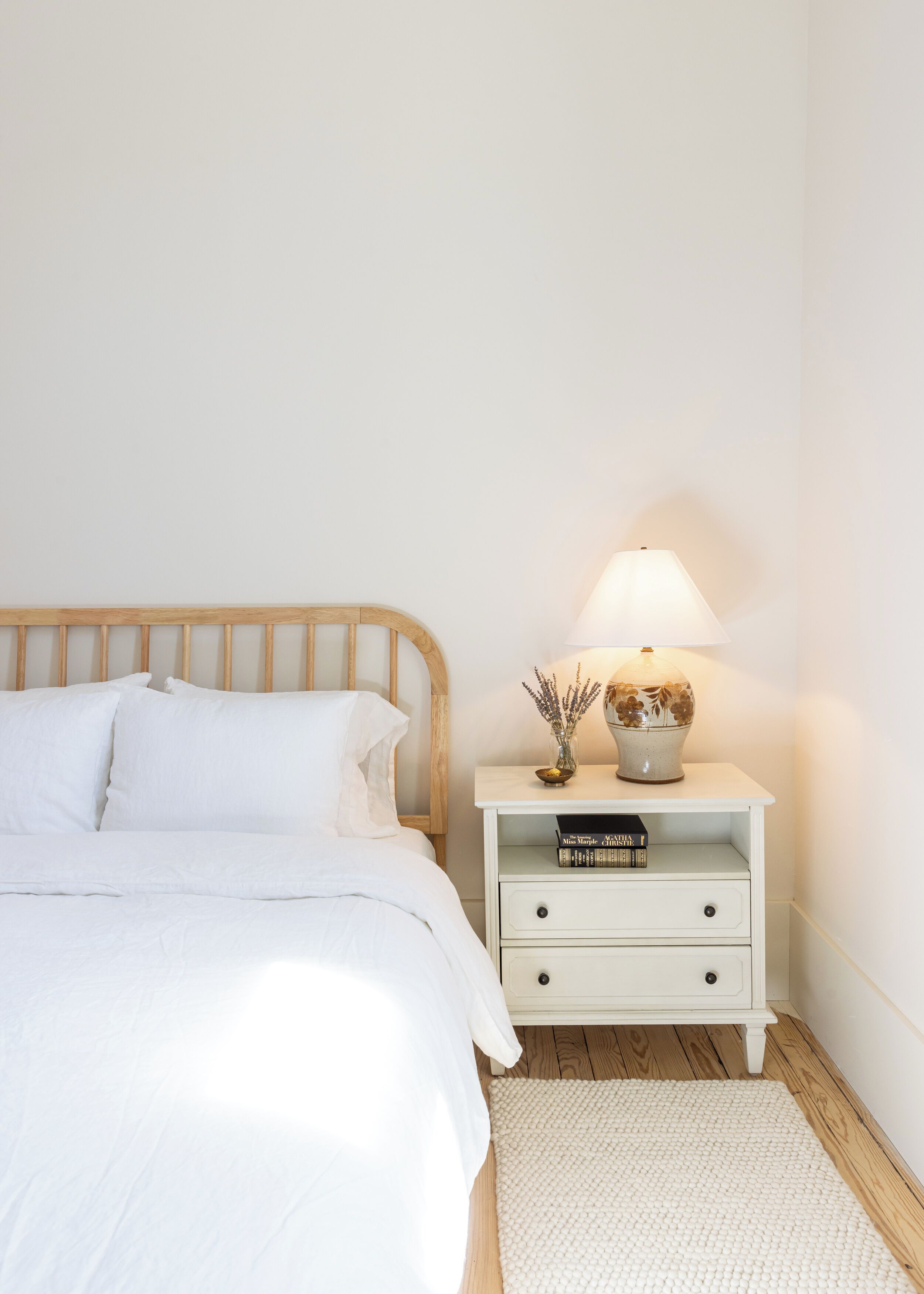 A neatly made bed with white bedding, a wooden headboard, a bedside table with a lamp, a vase with flowers, and a book titled "Miss Maple Christy".