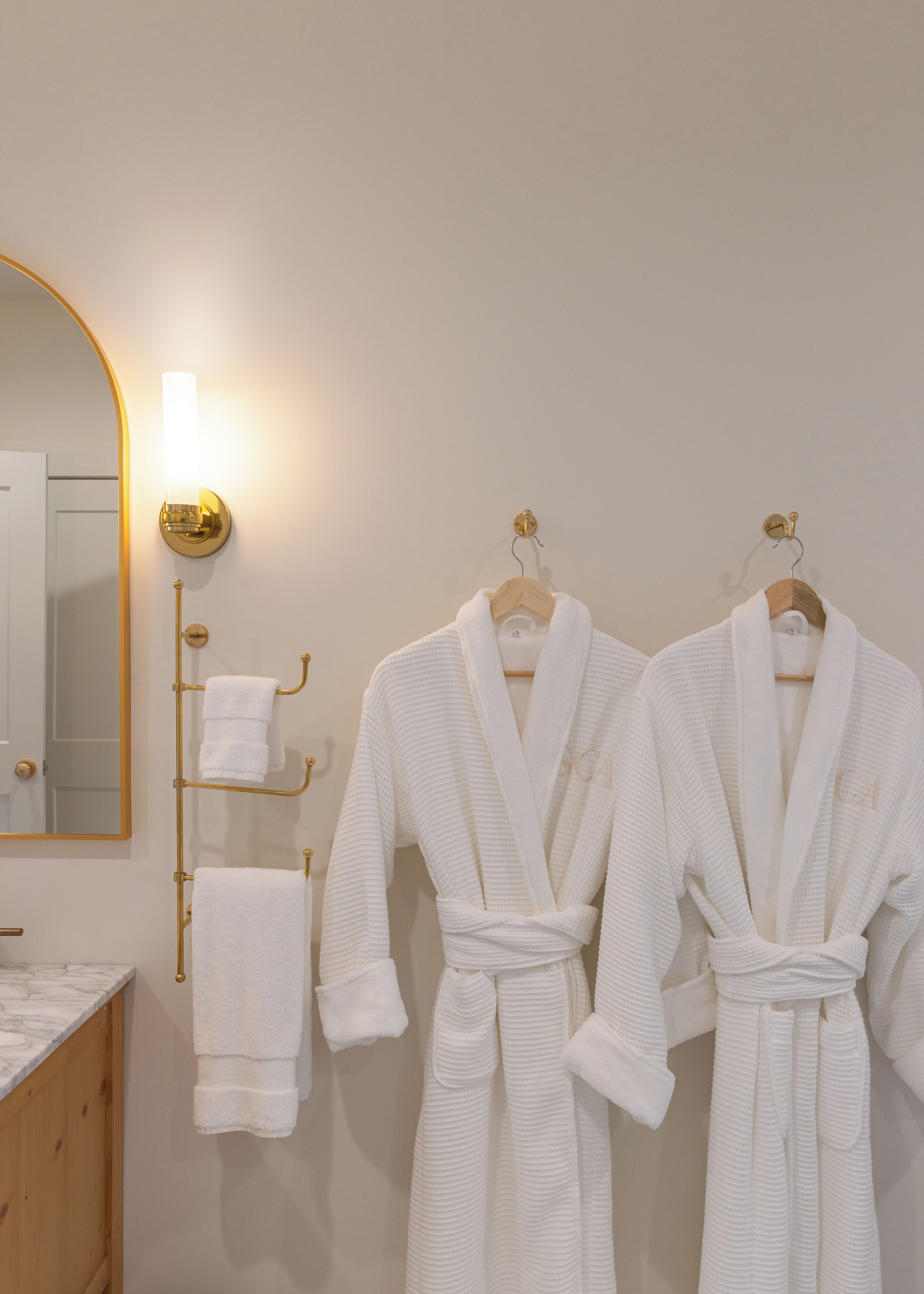 Ein Badezimmer mit zwei weißen Bademänteln, die an Holzaufhänger hängen, einem goldfarbenen Spiegel und einem Handtuchhalter mit weißen Handtüchern.