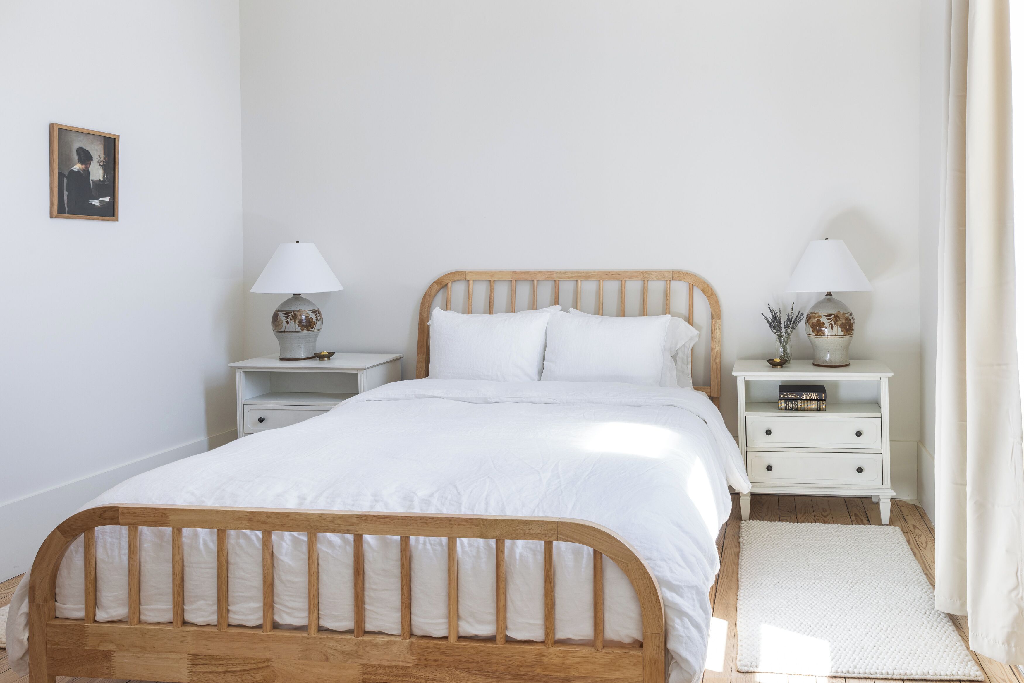Un dormitorio ordenado con cama de madera, ropa de cama blanca, dos mesitas de noche con lámparas y un pequeño tocador con un libro.