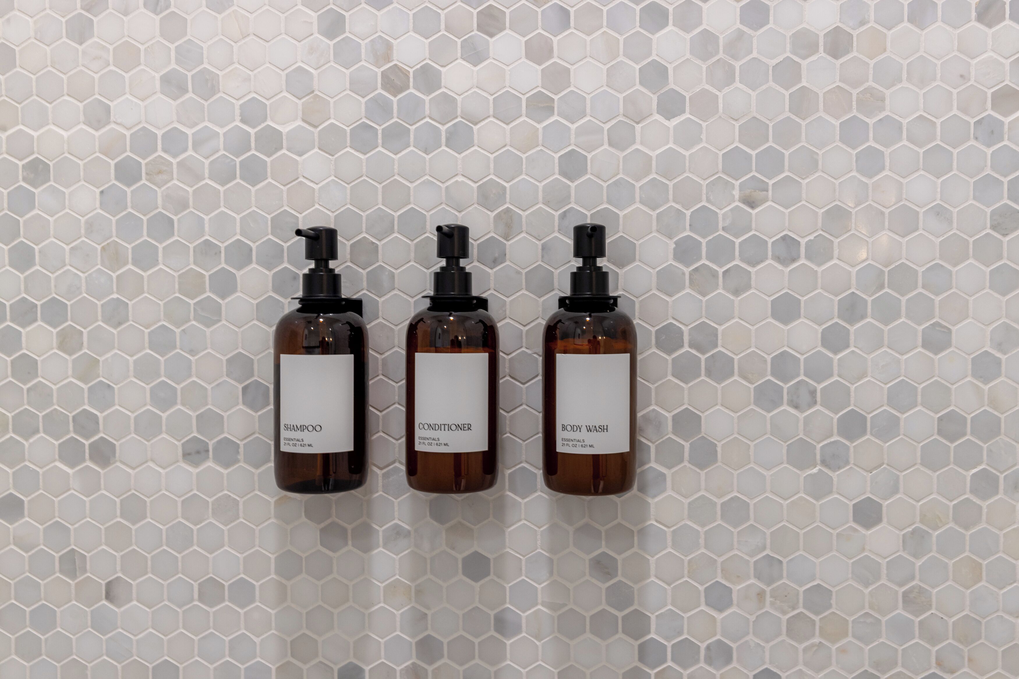 Três frascos de shampoo, condicionador e banho corporal em frente a uma parede de azulejos hexagonais.