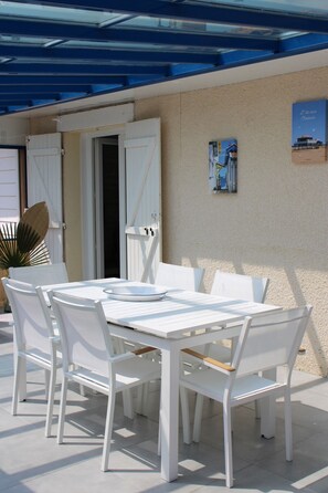 Un espace extérieur aménagé pour les repas, avec une table et des chaises, un coin salon abrité et un mur orné d’une affiche.
