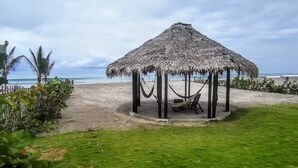 Un belvédère au toit de chaume, doté d’un hamac, entouré d’une pelouse et de palmiers, et offrant une vue sur la plage.
