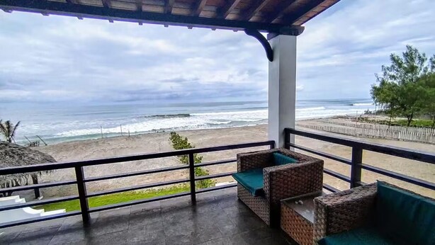 Un balcon abrité, doté de meubles en osier, offrant une vue sur la plage et surplombant un ciel nuageux.