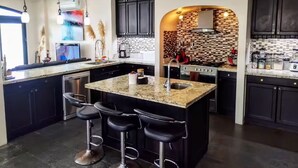 Une cuisine moderne avec des meubles de couleur sombre, un îlot central en granit doté de tabourets, et un pare-feu en carreaux.