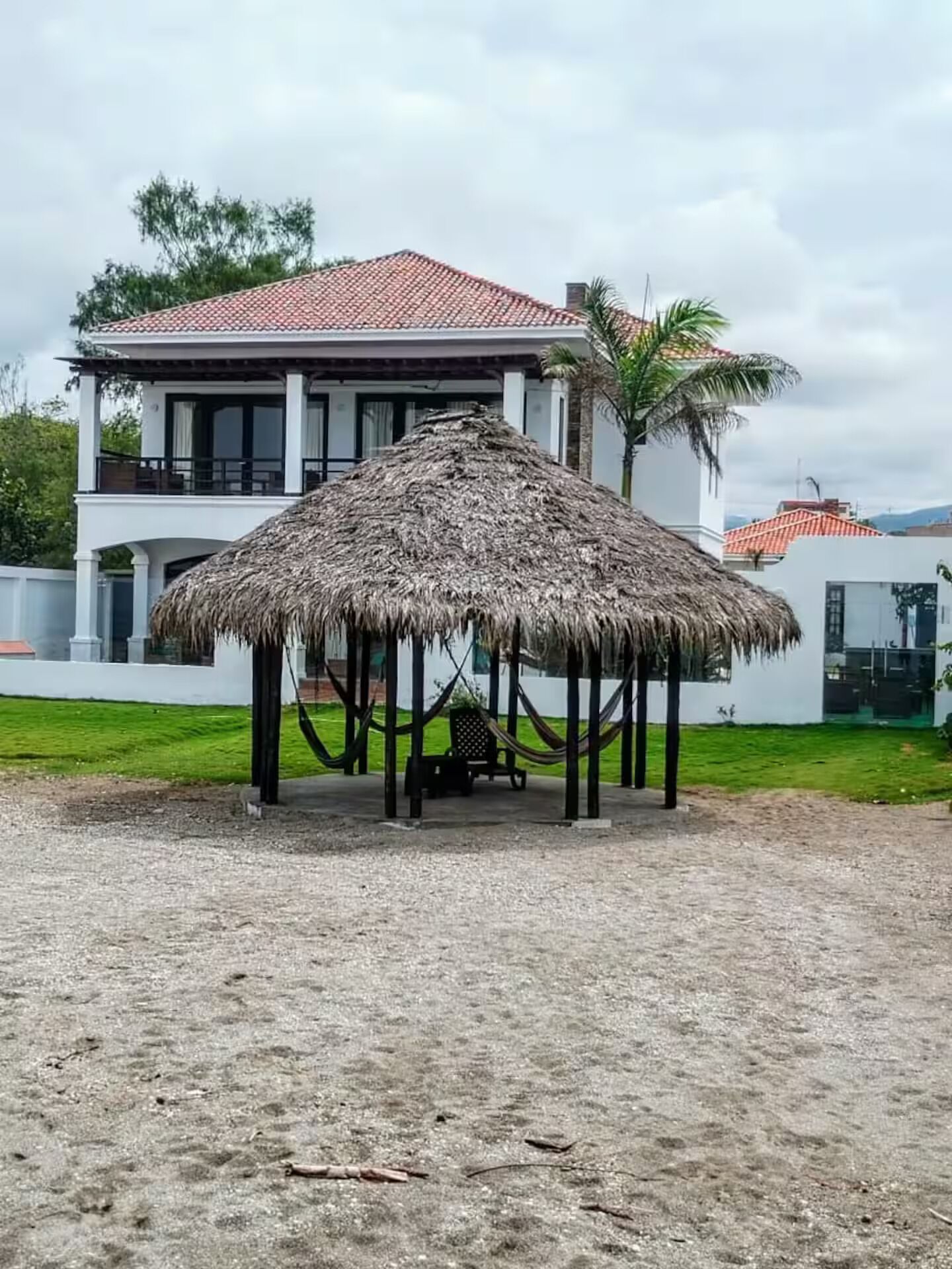 A thatched-roof gazebo in a sandy area with a white building in the background.