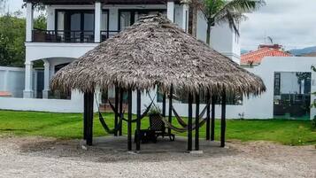 A thatched-roof gazebo in a sandy area with a white building in the background.