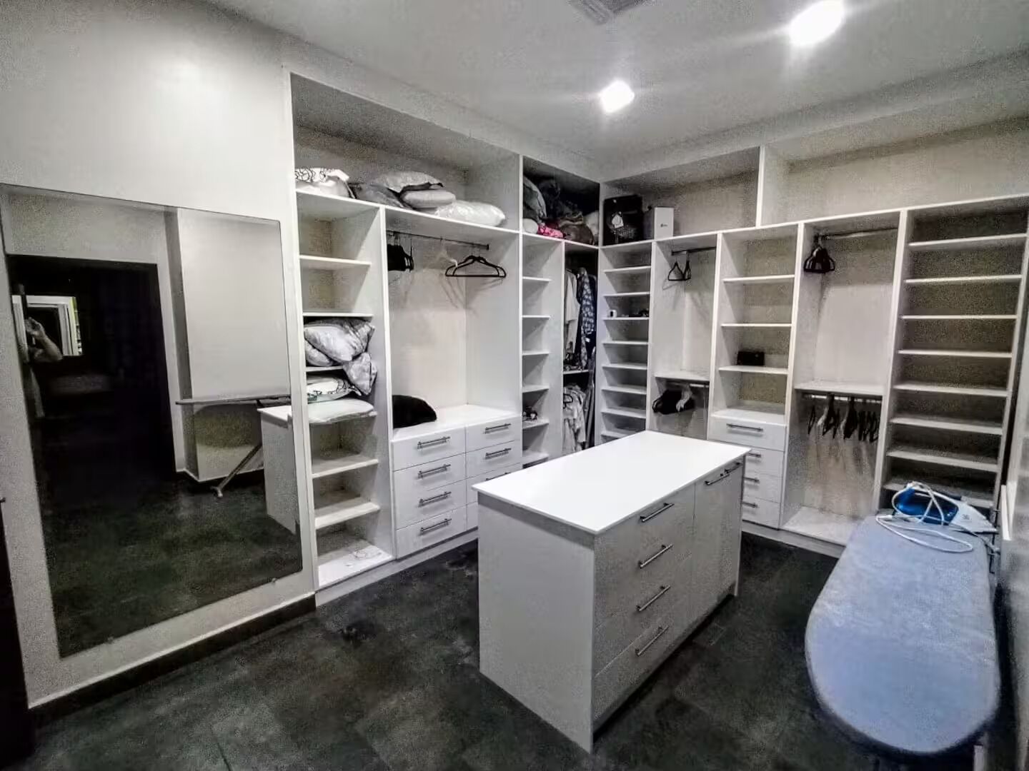 Ein begehbarer Kleiderschrank mit zahlreichen Regalen, Schubladen und Ablageflächen zum Aufhängen, mit einer zentralen weißen Kommode und einer gebogenen Sitzbank.