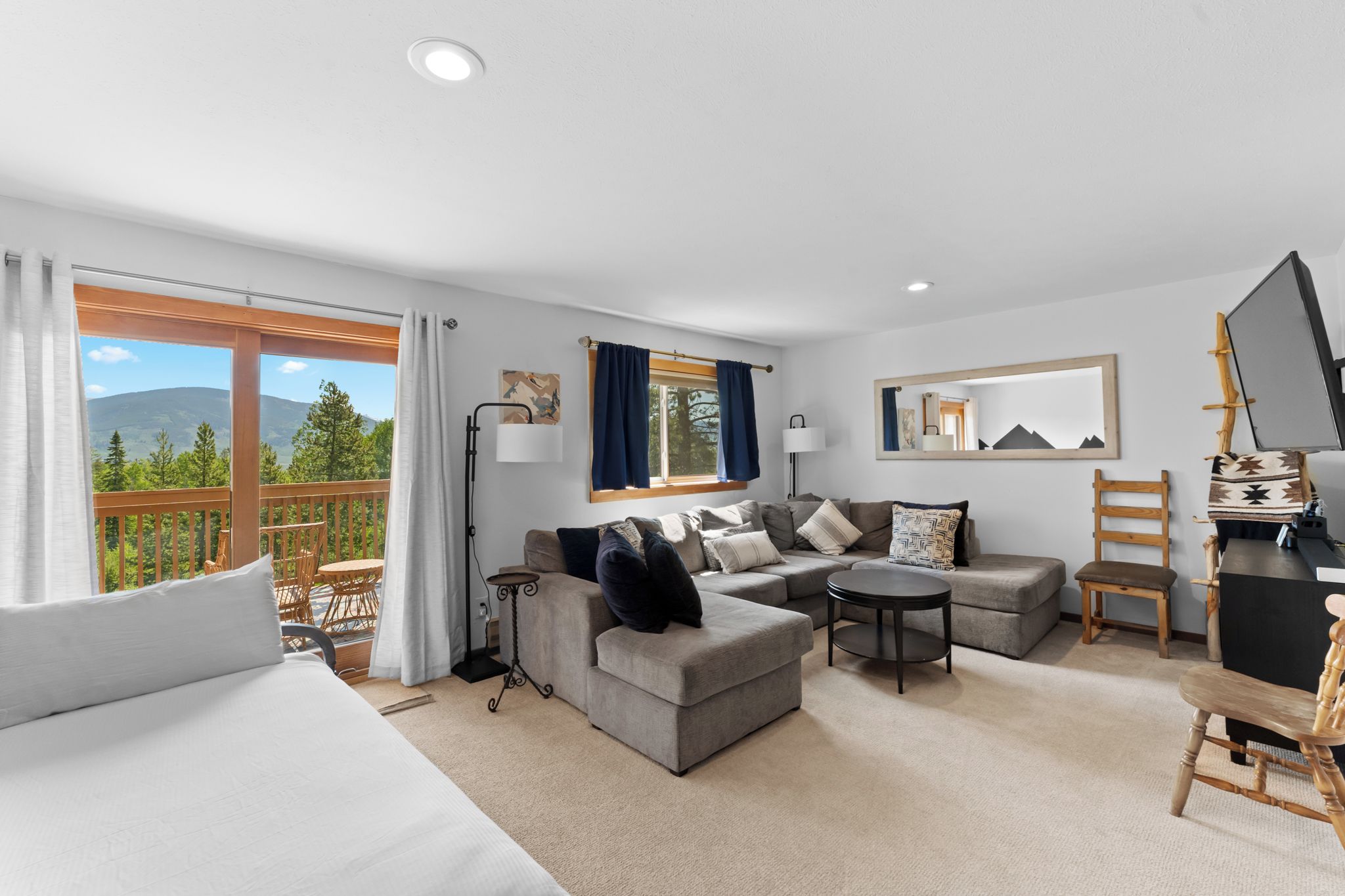 A modern hotel room with a large bed, a sofa, a small table, and a balcony with a view of mountains.