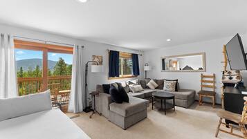 A modern hotel room with a large bed, a sofa, a small table, and a balcony with a view of mountains.