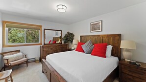 Een slaapkamer met een groot bed, houten hoofdeinde, nachtkastjes, een bankje, een raam met uitzicht op groen en een plafondlamp.