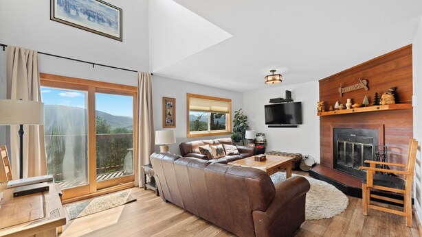 Een ruime woonkamer met een bruine leren bank, een houten salontafel en een open haard. Er is een groot raam met uitzicht op de omgeving, en een televisie aan de wand bevestigd.