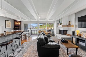 Een ruime woonkamer met een grote bank, een houten salontafel en een open haard. Er is een keuken met een bar en barkrukken. De kamer heeft grote ramen met uitzicht op de omgeving.