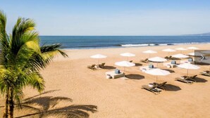 A beach with white umbrellas and lounge chairs, palm trees, and a clear view of the ocean.