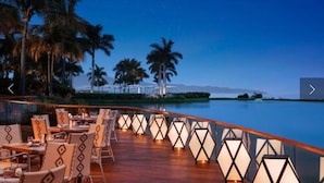 A waterfront restaurant with outdoor seating, palm trees, and a view of the ocean.