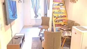 Un salon compact comprenant une télévision, deux fauteuils en osier, une bibliothèque remplie de jouets et un petit réfrigérateur.
