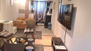 Un salon compact comprenant un canapé, des fauteuils, une petite table, un téléviseur et, au fond, un coin cuisine.