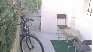 Un vélo appuyé contre un mur, une petite table avec des chaises et un tapis vert sur le sol.