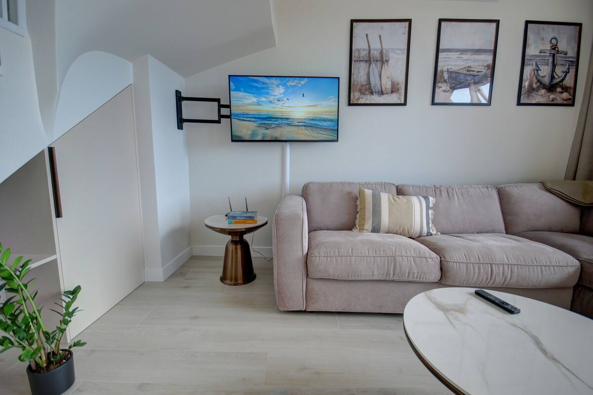 A living room with a beige sofa, a round coffee table, a television mounted on the wall, and a small side table with books.