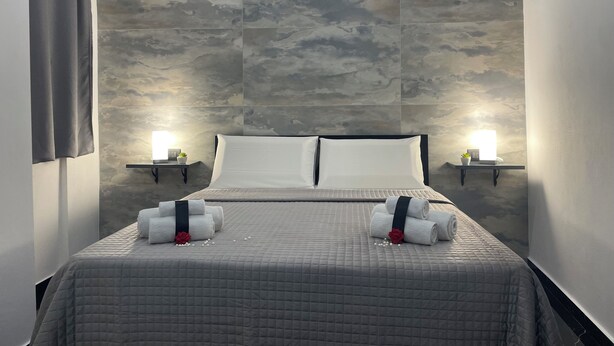 Una cama bien hecha con una colcha gris acolchada, dos toallas enrolladas con una rosa roja, y una mesita de noche con una lámpara y una planta en maceta.