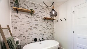 A bathroom with a white bathtub, a brick-patterned wall, wooden shelves, a hanging light fixture, and a white door. - Aux Repères de Papi (Lac-Etchemin)