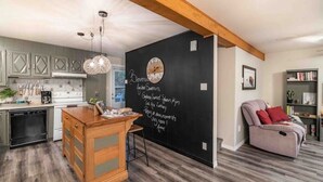 A modern kitchen with a chalkboard wall, wooden cabinets, and a dining area with a cushioned chair. - Aux Souvenirs doux CITQ 303027 (Lac-Etchemin)