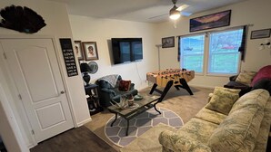 A living room with a sofa, a coffee table, a foosball table, a ceiling fan, and a window with blinds.
