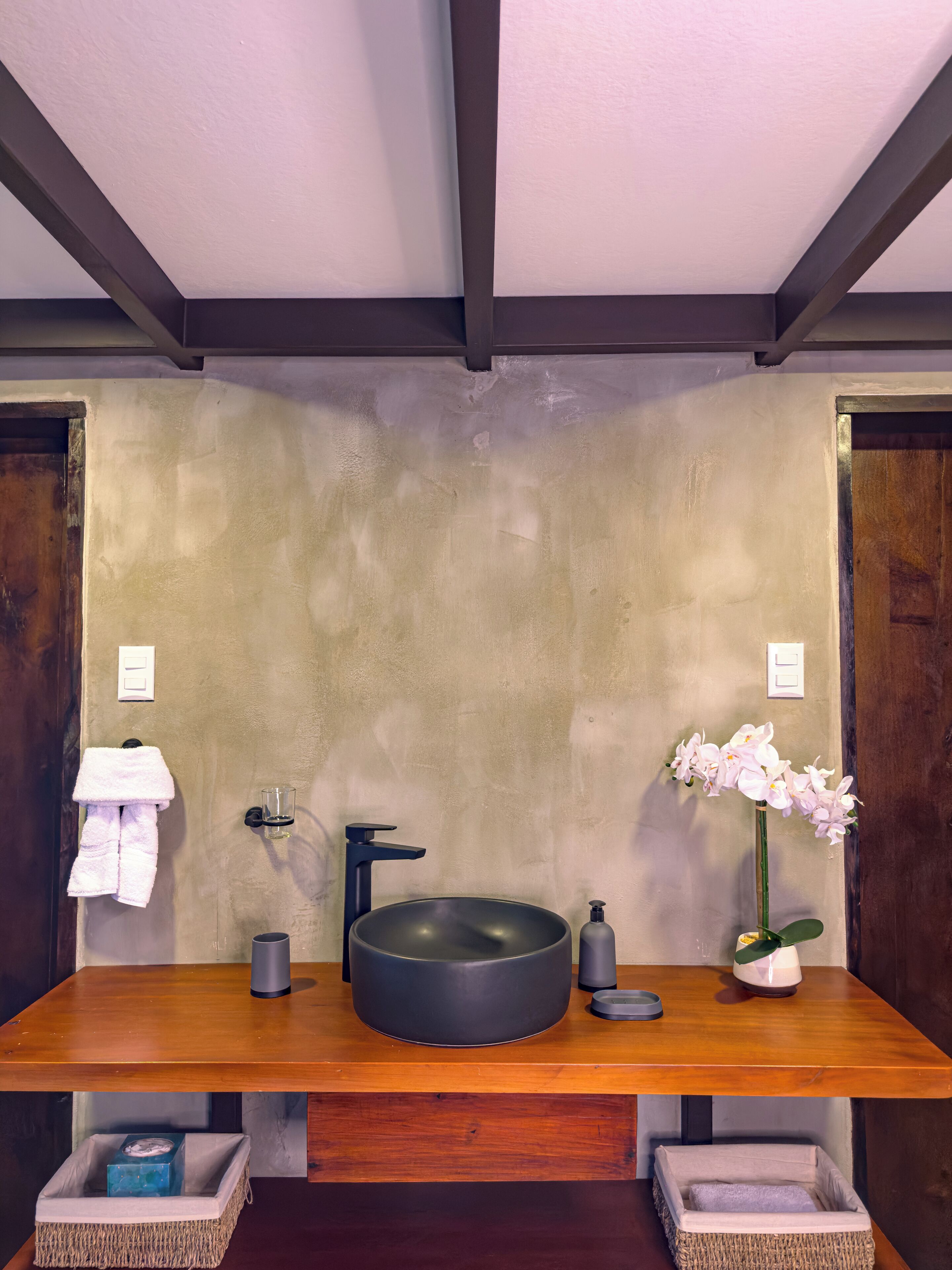 Un baño con un lavabo negro redondo, un perchero con una toalla blanca, un vaso de agua, una orquídea en maceta y dos cestas de mimbre.