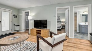 Un salon moderne doté d’un téléviseur à écran plat, d’un canapé, d’une table basse et d’un coin cuisine visible à travers une porte ouverte.