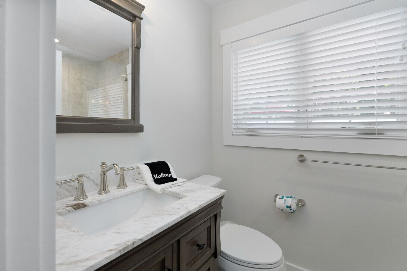A bathroom with a white vanity, a mirror, a towel rack with a 'Makeup' towel, a toilet, and a window with blinds.