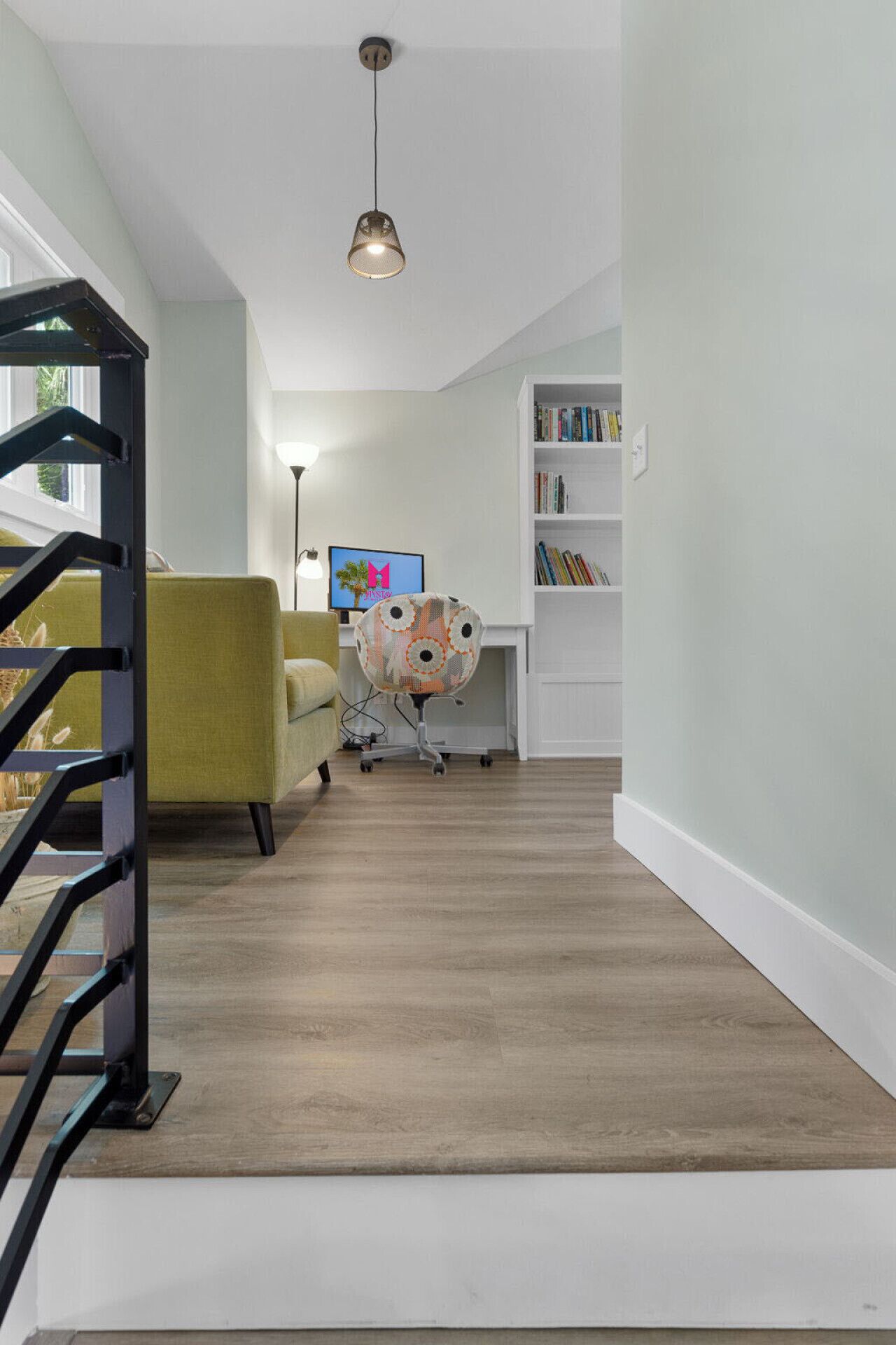 A modern living room with a staircase, a yellow armchair, a desk with a computer, and a bookshelf.