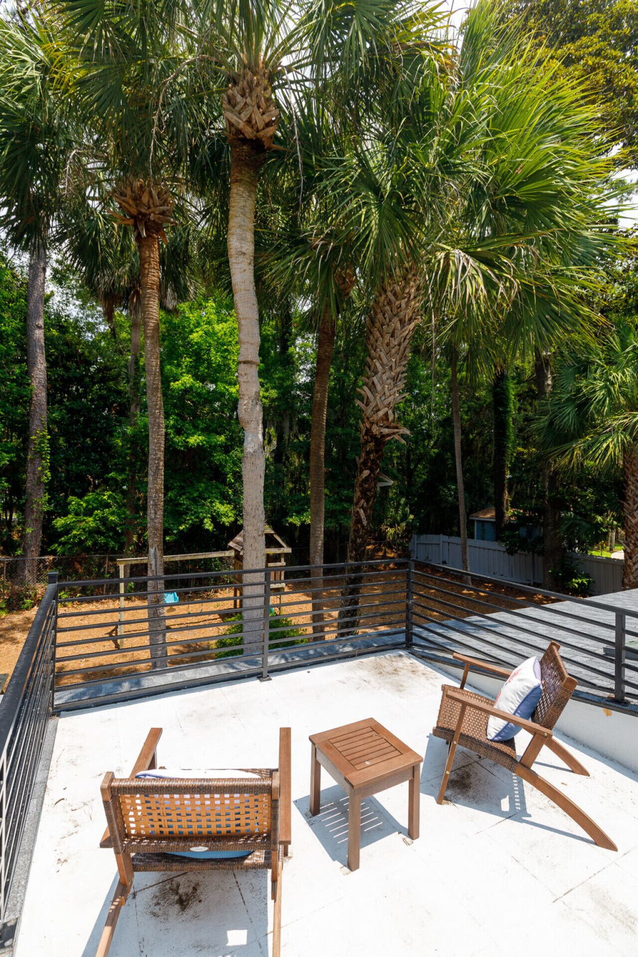 Una terraza en la azotea con un banco de madera, una mesita y una silla, rodeada de altas palmeras y una barandilla de metal.