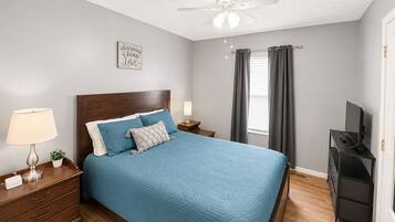 Une chambre à coucher comprenant un lit, une table de chevet, une lampe, un ventilateur de plafond, un téléviseur et une fenêtre avec des rideaux.