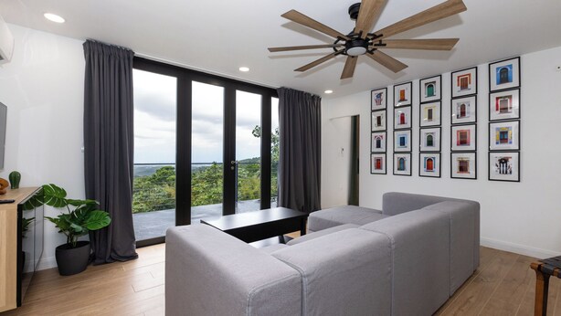 Uma sala de estar moderna com um sofá seccional cinza, um ventilador de teto de madeira e uma janela grande com cortinas.