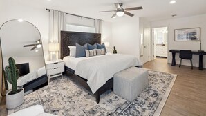 A modern bedroom with a large bed, a ceiling fan, a nightstand, a cactus, and a desk with a chair.