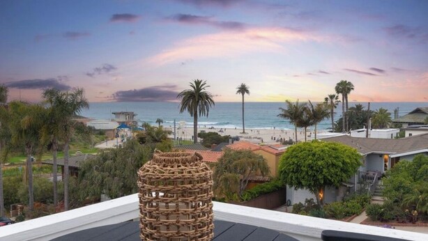 Vista costera desde un balcón con vista a una playa, palmeras y un atardecer.