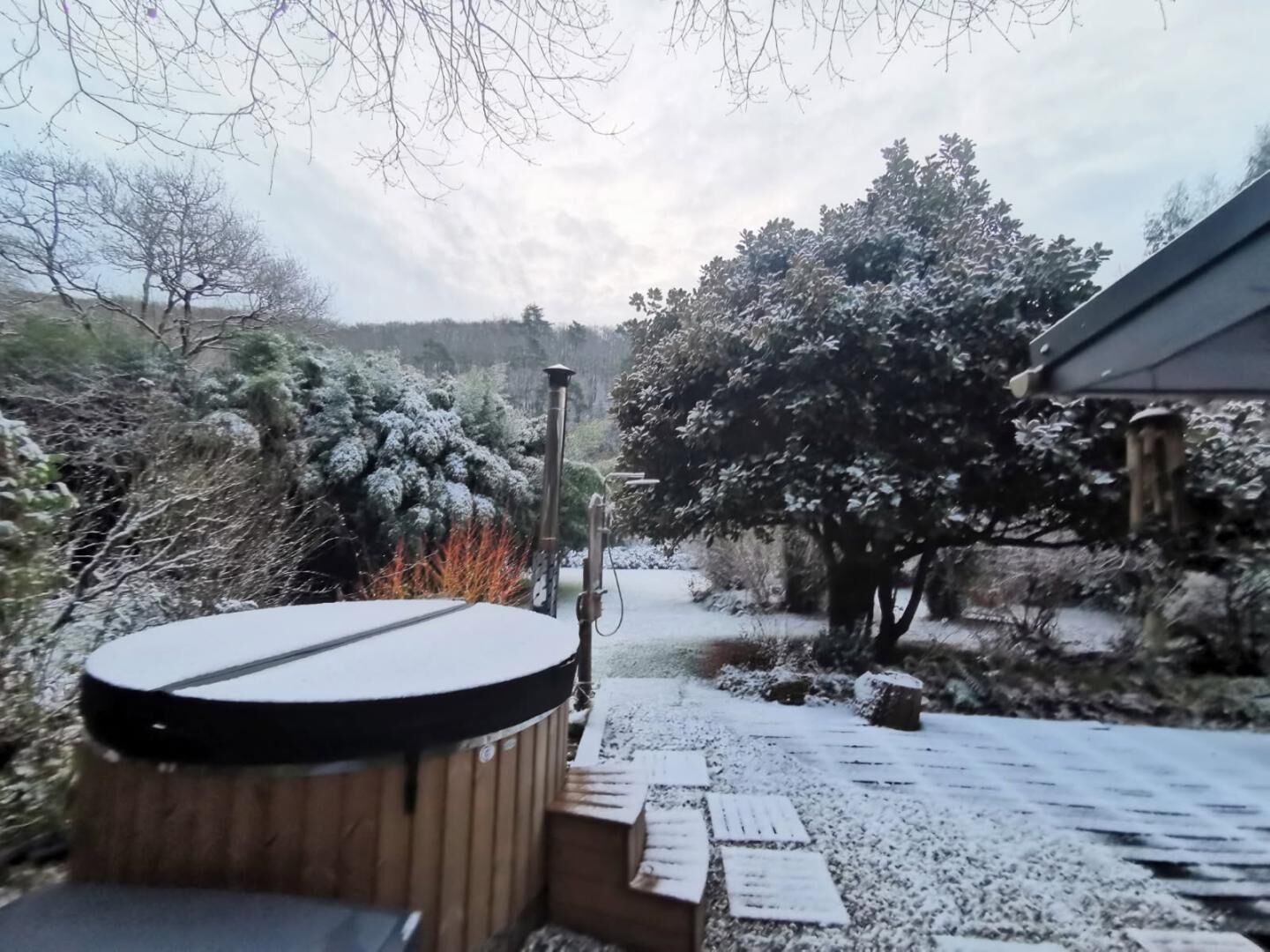 Een houten terras met een jacuzzi, omgeven door met sneeuw bedekte bomen en een stenen pad.