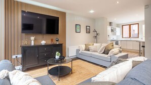 Una sala moderna con televisor de pantalla plana, un mueble de madera oscura, un sofá gris y una cocina al fondo.