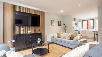 A modern living room with a flat-screen TV, a dark wooden cabinet, a grey sofa, and a kitchen area in the background.