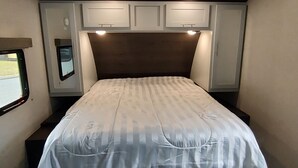 A bedroom with a bed, overhead cabinets, and a window. - Cozy RV in Quaint Klamath with AC, WiFi, DISH network, fully hooked up. (Klamath)