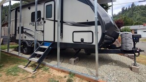 A white recreational vehicle with a covered living area, a ladder, and a water heater. - Cozy RV in Quaint Klamath with AC, WiFi, DISH network, fully hooked up. (Klamath)