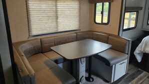 A compact RV interior with a dining area, cushioned seating, a small table, and a window with blinds. - Cozy RV in Quaint Klamath with AC, WiFi, DISH network, fully hooked up. (Klamath)