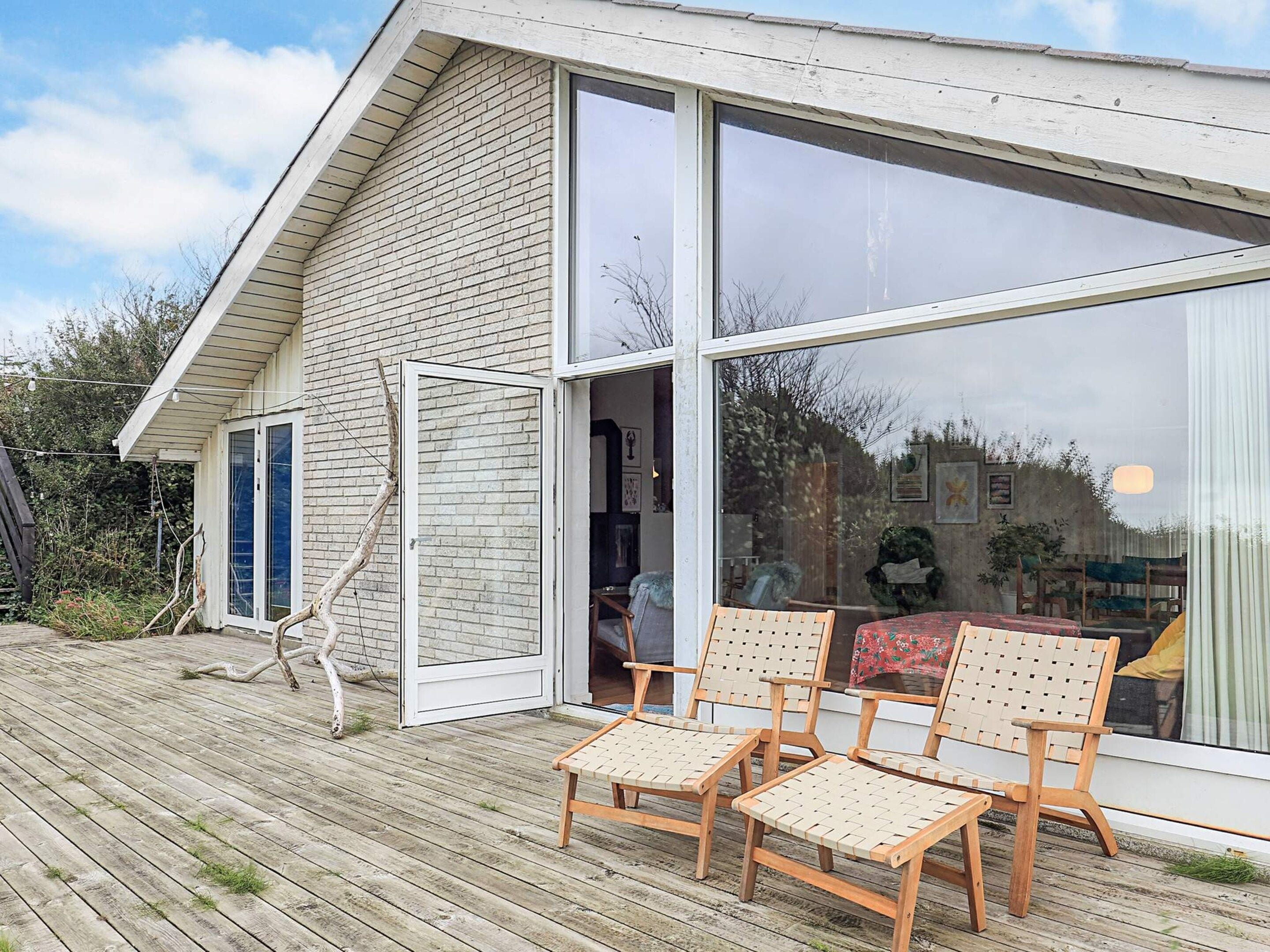 Uma casa moderna com uma grande janela de vidro, um deck de madeira com cadeiras e uma porta que dá para o interior.