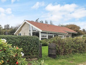 Ein eingeschossiges Haus mit geneigtem Dach, umgeben von dichtem Buschwerk und einem Holztor.