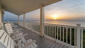Een overdekt terras met witte leuningen en stoelen gericht op de oceaan bij zonsondergang.