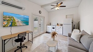 A compact living space with a kitchenette, a dining area with a desk and chair, a sofa, a ceiling fan, and a large TV displaying a beach scene. - The Seapearl at UTC-1/1 Guest Suite (Sarasota)