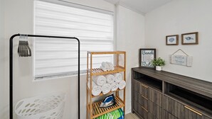 A laundry area with a rack holding towels, a wooden shelf with rolled towels, and a white chair. - The Seapearl at UTC-1/1 Guest Suite (Sarasota)
