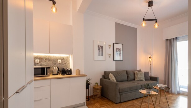 Een moderne keuken met witte kasten, een magnetron en een koffieapparaat. Er staat een grijze bank met kussens en een kleine ronde tafel met een plant. De kamer heeft een houten vloer en een groot raam met gordijnen.