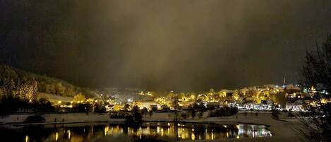 A nighttime view of a town with buildings illuminated by street lights, a body of water reflecting the lights, and snow on the ground.