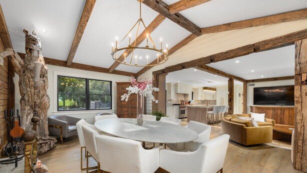 A spacious living area with a round dining table, white chairs, a chandelier, and wooden beams on the ceiling.